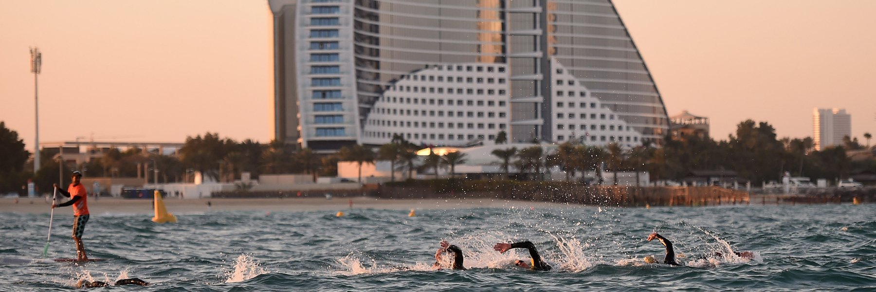 IRONMAN 70.3 Dubai athletes are swimming in the Arabian Gulf next to the Burj al Arab while a volunteer keeps watching them on a surf board at sunrise