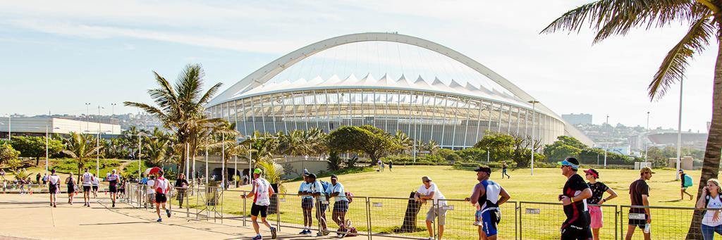 Athletes are running in the park along a palm tree avenue and passing by Moses Mabhida Stadium while supporters are watching and cheering on at IRONMAN 70.3 Durban