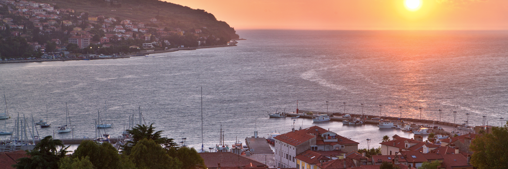 Two opposite cities that are separated by the sea at sunset, which turns the sky and sea a little reddish in Slovenia