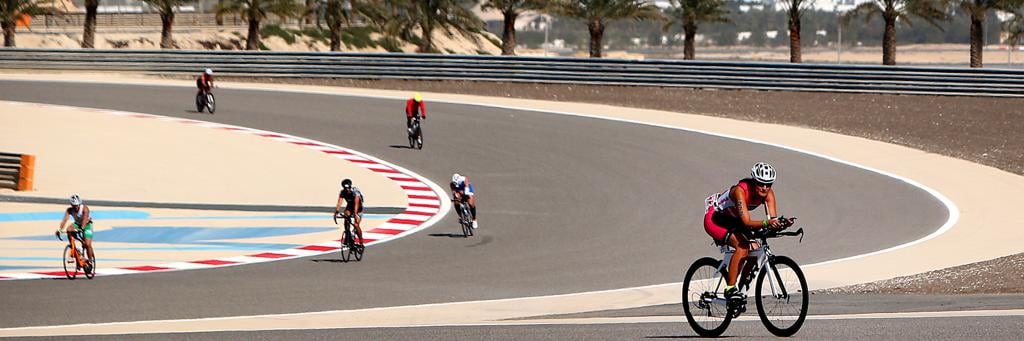 Athletes are biking through the for motorsport used venue Bahrain International Circuit which is lined with palm trees and sand