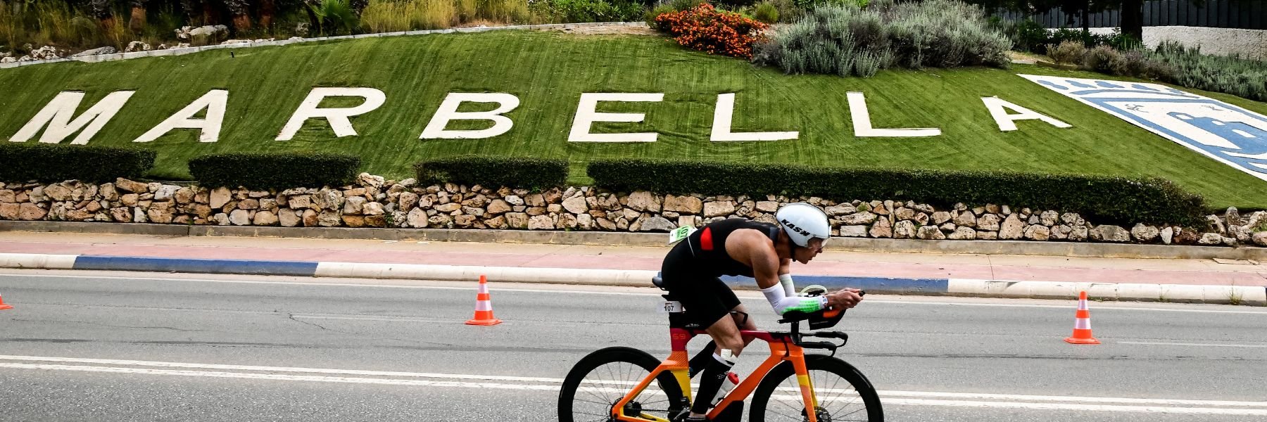 Two athletes biking on a street with a small town of white houses located on a small green hill in the background at IRONMAN 70.3 Marbella
