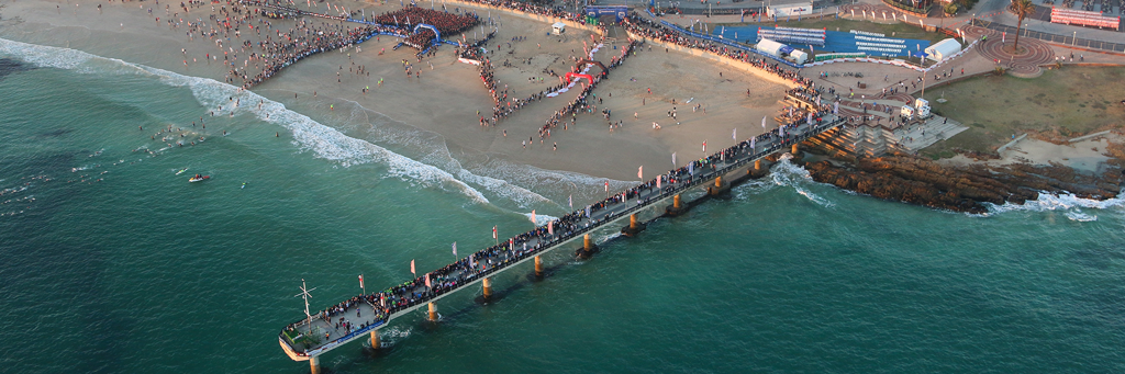 Scenic image from above of swim start at Hobie Beach at Nelson Mandela Bay / Port Elizabeth with athletes swimming or waiting to get into the sea and people watching at a pier