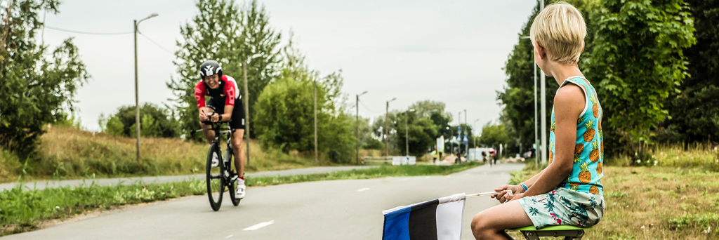 Little boy waving with the Estonian flag while a smiling athlete is passing by on his bike 