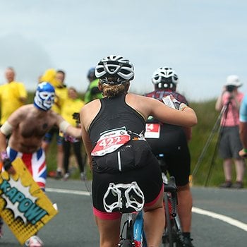 Athletes who are biking up a hill are motivated and supported by masked people carrying posters at IRONMAN UK